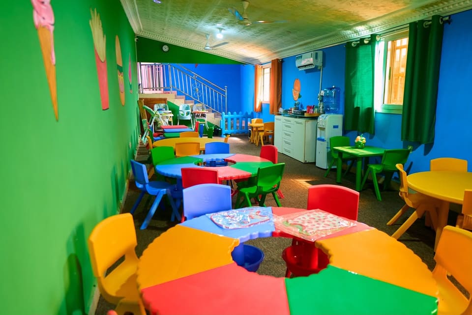 Une salle d’activités aux murs verts, avec des tables rondes multicolores et des chaises à hauteur d’enfant.