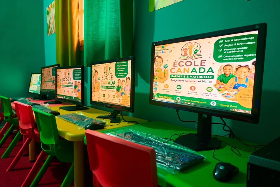 La salle d’informatique : une rangée d’écrans posés sur un plan de travail vert, avec des chaises d’enfant de couleurs vives.